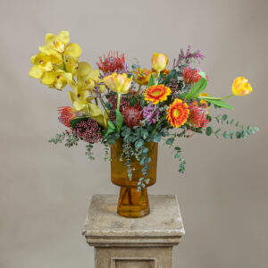 Arreglo Tropical Mom con protea pincushion, cymbidium, gerberas y tulipanes en florero de vidrio estilo cáliz, ideal para el Día de las Madrea